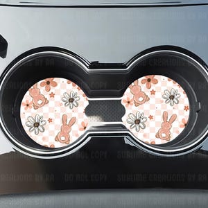May include: Two car coasters with a pink and white floral and bunny design. The coasters feature a checkered background with daisies and bunnies. The coasters are designed to fit in a car's cup holders.