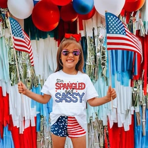 Op de afbeelding: Een kind met een zonnebril, een wit t-shirt met de tekst "Star Spangled and Sassy" en een patriottische korte broek. Ze houdt Amerikaanse vlaggen vast voor een achtergrond van rood, wit en blauwe decoraties, waaronder ballonnen en slingers.