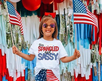 Puede incluir: Una joven con gafas de sol, una camiseta blanca con el texto "Star Spangled and Sassy" y pantalones cortos patrióticos. Sostiene banderas americanas frente a un telón de fondo de decoraciones rojas, blancas y azules, incluyendo globos y serpentinas.