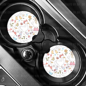 May include: Two car coasters with a white background featuring a pattern of cartoon geese, flowers, butterflies, and boots. The coasters are placed in a car's cup holders, with a black and silver dashboard visible.