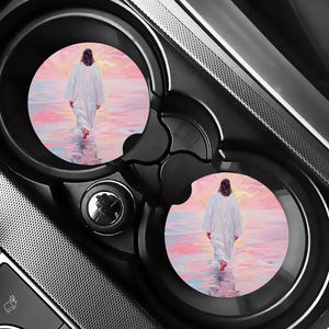 May include: Two circular car cup holder coasters featuring a printed illustration of a figure in a long white robe walking across a calm body of water toward a soft pink and orange sunset sky, with reflections visible on the water surface.