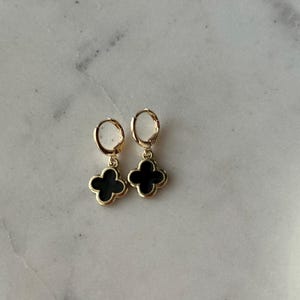 May include: A pair of gold hoop earrings with black clover-shaped charms. The earrings are simple and elegant.