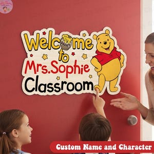 Op de afbeelding: Een houten klaslokaalbord met Winnie de Poeh, met de tekst "Welkom in de klas van mevrouw Sophie". Het bord heeft gele, rode en zwarte letters en een cartoonillustratie van Winnie de Poeh. Het bord staat op een rode deur.