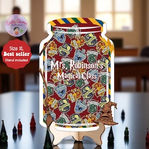 Könnte beinhalten: Ein dekoratives, gläserförmiges Schild mit dem Text "Mrs. Robinson's Magical Class" und verschiedenen Hauswappen. Das Schild enthält einen Holzständer, einen Besen und einen Hut. Der Hintergrund ist ein verschwommenes Interieur.