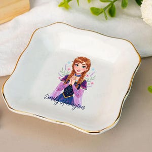 May include: White ceramic ring dish with a gold trim and a cartoon illustration of a princess. The dish has a wavy edge and the name "Emily Harrington" is printed on the dish. A silver ring with a large stone is next to the dish.