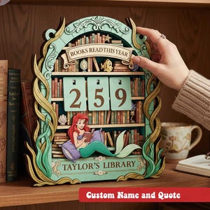 May include: A wooden calendar shaped like a bookshelf, featuring the text "BOOKS READ THIS YEAR" and the numbers "259". The design includes a mermaid reading a book, with the text "TAYLOR'S LIBRARY". The calendar is displayed on a wooden shelf.