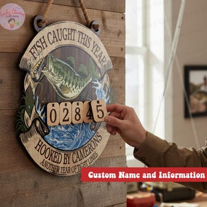May include: Wooden fishing sign with the text "FISH CAUGHT THIS YEAR" and "HOOKED BY CAMERONS". The sign features a fish graphic and a space to display the number of fish caught. Includes a rope for hanging.