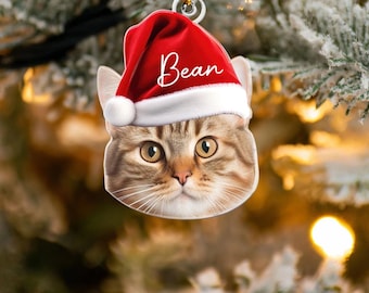 May include: A cat-shaped Christmas ornament featuring a tabby cat wearing a red Santa hat with the name "Bean" in white script. The ornament has a gold string for hanging and is set against a blurred background of a Christmas tree.