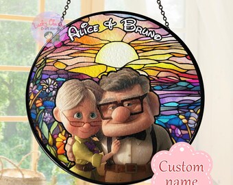May include: A stained glass style round window hanging with a black frame. The image features a cartoon couple, a man and a woman, with a sunset background. The text "Alice & Bruno" is written in a decorative font at the top of the image.