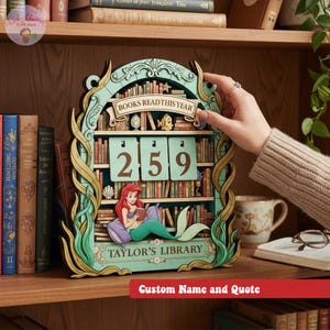 May include: A wooden bookshelf-shaped calendar with the text "BOOKS READ THIS YEAR" and the numbers "259". The design includes a mermaid reading a book, with the text "TAYLOR'S LIBRARY". The calendar is on a wooden shelf.
