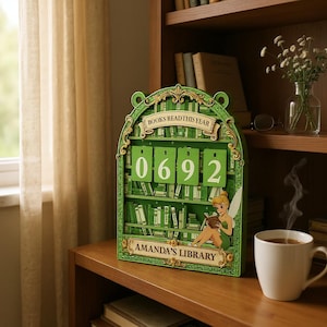 May include: A green wooden book counter with the text "BOOKS READ THIS YEAR" and the numbers "0692". The counter has a bookshelf design with a Tinkerbell illustration and the text "AMANDA'S LIBRARY". A white mug of coffee sits nearby.
