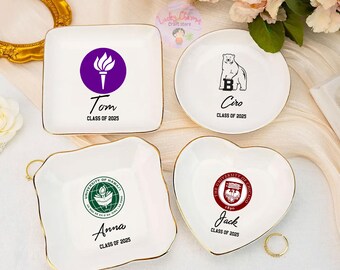 Puede incluir: Cuatro platos de anillo de cerámica blanca con ribete dorado. Cada plato presenta un logotipo de universidad diferente y el nombre de un graduado. Los logotipos son para la Universidad de Hawái, la Universidad de Chicago, la Universidad de Boston y la Universidad de Alaska. Los platos están etiquetados con los nombres Anna, Jack, Ciro y Tom, y el año 2025.