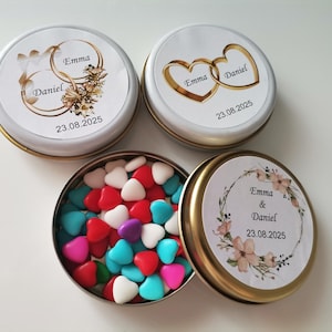 Puede incluir: Pequeñas latas redondas con borde dorado y tapas blancas, cada una con un diseño personalizado para una boda. Una lata está abierta, revelando caramelos coloridos en forma de corazón. Las tapas muestran nombres y una fecha.