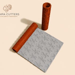 Puede incluir: Un juego de herramientas para arcilla polimérica, que incluye un rodillo texturizado y una lámina estampada. El rodillo es de color marrón rojizo con un diseño floral. La lámina es gris y presenta un patrón de libélulas. La marca "Pampa Cutters" es visible.
