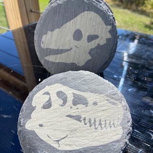 May include: A stack of gray slate coasters, each featuring a cream-colored dinosaur skull design. The coasters are round and have a natural, textured surface, perfect for adding a touch of prehistoric flair to any home decor.