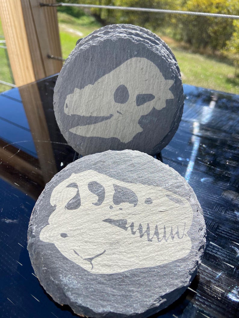 May include: A stack of gray slate coasters, each featuring a cream-colored dinosaur skull design. The coasters are round and have a natural, textured surface, perfect for adding a touch of prehistoric flair to any home decor.