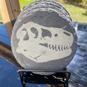 May include: A set of round, dark gray slate coasters. Each coaster features a white dinosaur skull design. The coasters are stacked on a black metal stand, suitable for home decor and protecting surfaces from drink rings.