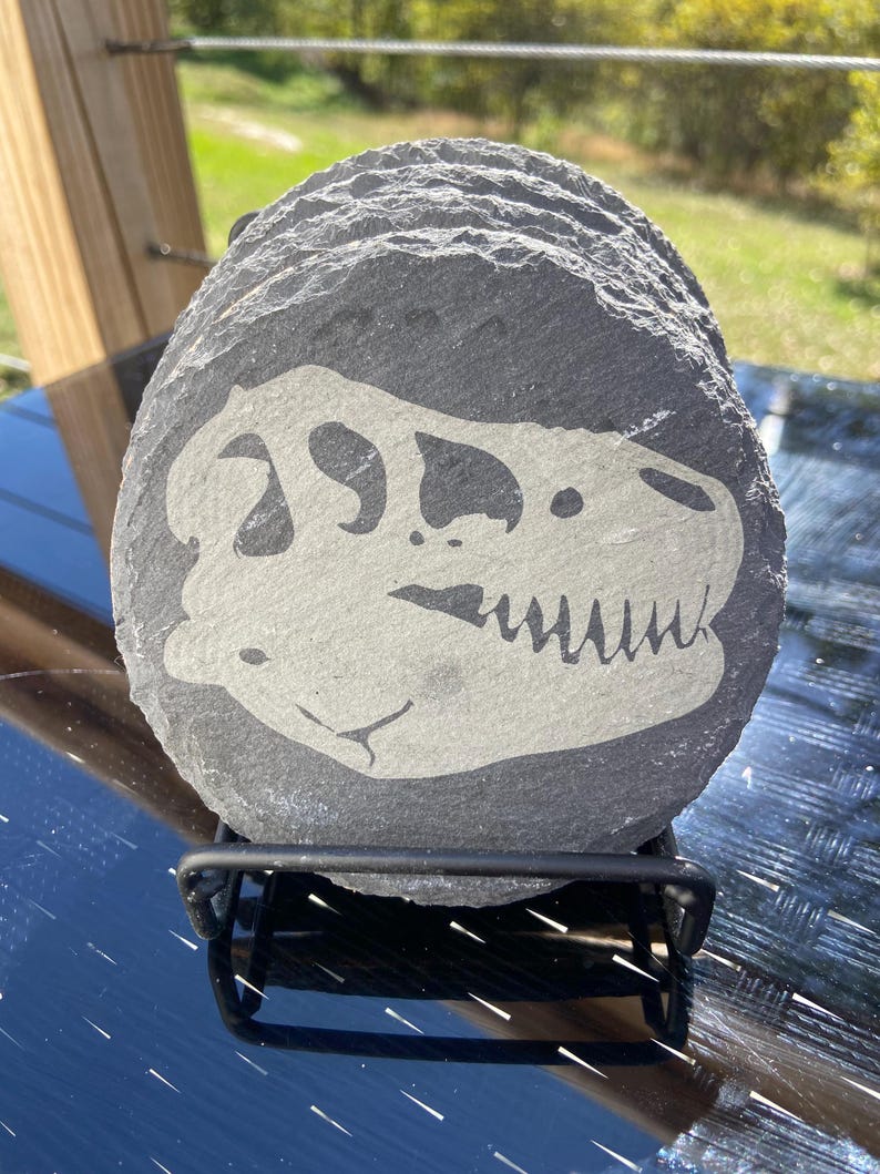 May include: A set of round, dark gray slate coasters. Each coaster features a white dinosaur skull design. The coasters are stacked on a black metal stand, suitable for home decor and protecting surfaces from drink rings.
