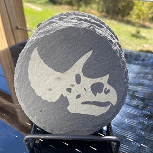 May include: A stack of gray, round slate coasters. Each coaster features a white graphic of a Triceratops skull. The coasters are displayed on a black metal rack, perfect for home decor.