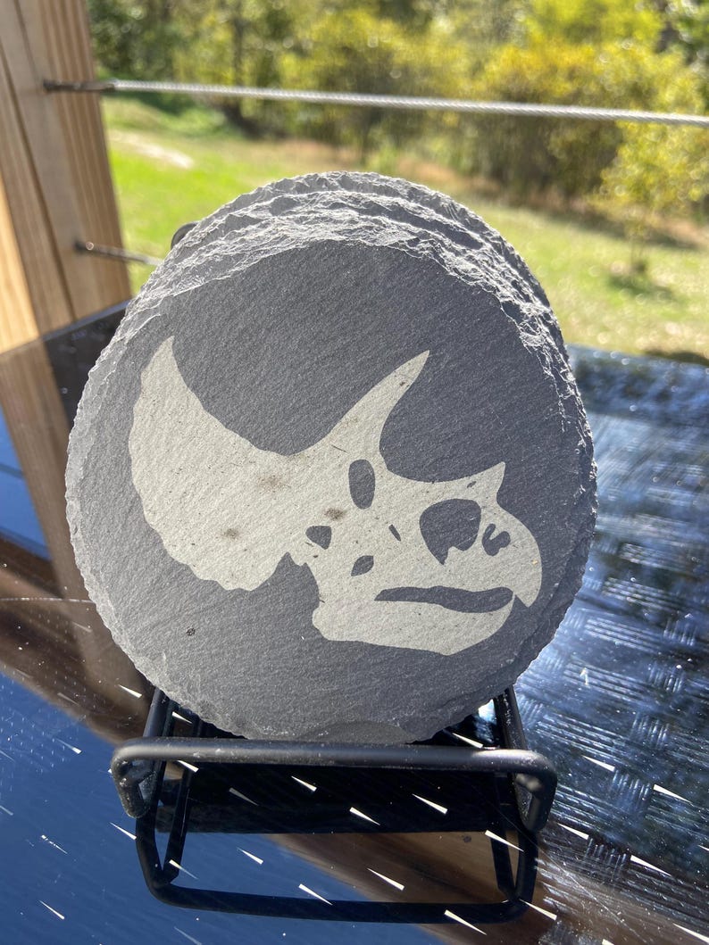 May include: A stack of gray, round slate coasters. Each coaster features a white graphic of a Triceratops skull. The coasters are displayed on a black metal rack, perfect for home decor.