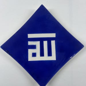 Puede incluir: Un azulejo de cerámica azul con un diseño de caligrafía árabe blanca. El azulejo tiene forma de diamante.