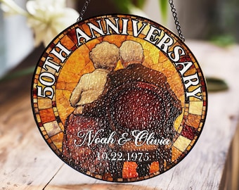 May include: A round stained glass window hanging from a chain. The window is decorated with a mosaic pattern in shades of orange, yellow, and brown. The text "50th Anniversary" is written in a white circle around the edge of the window. The centre of the window features a couple sitting together, with the text "Noah & Olivia 10.22.1975" below them.