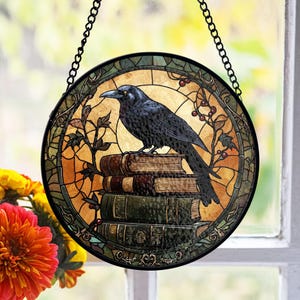 May include: A stained glass window hanging features a black raven perched on a stack of old books. The raven has yellow eyes and is surrounded by a colorful stained glass design with vines and berries.