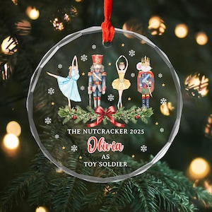 May include: A clear, round Christmas ornament with a red ribbon. The ornament features illustrations of characters from The Nutcracker ballet, including a ballerina, a nutcracker, and a mouse king. Text reads "THE NUTCRACKER 2025" and "Olivia AS TOY SOLDIER".