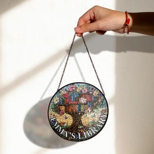 May include: A round, stained glass window hanging ornament featuring a tree with books for leaves. The tree is surrounded by flowers and butterflies. The text "Emma's Library" is written around the bottom of the ornament.