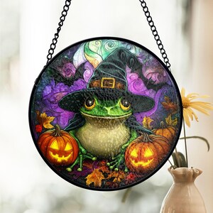May include: Round Halloween-themed hanging decoration featuring a green frog wearing a witch hat. The frog is surrounded by pumpkins, bats, and autumn leaves, set against a purple and green background. A black chain is attached for hanging.