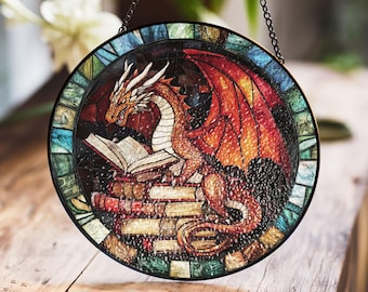 Peut inclure: Une fenêtre en verre coloré suspendue représente un dragon rouge lisant un livre sur une pile de livres. Le dragon a un corps vert et jaune avec des ailes rouges. L'arrière-plan est une mosaïque de verre coloré.