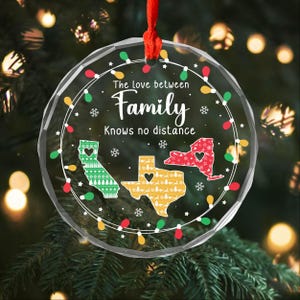 Op de afbeelding: Een heldere, ronde kerstversiering met een rood lint. De versiering toont de tekst "The love between Family Knows no distance" en contouren van Californië, Texas en New York. De staten zijn versierd met feestelijke patronen en een hart.