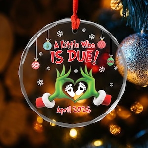 May include: A clear, round Christmas ornament with the text "A Little Who IS DUE!" in red. The ornament features green hands forming a heart around baby footprints, with the date "April 2026" below. Red ribbon for hanging.