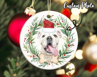 May include: A round Christmas ornament featuring a white bulldog wearing a Santa hat and a wreath of green leaves and red berries. The dog's name, "Charlie," and the year "2023" are printed on a bone-shaped tag. The words "Custom Photo" are written on the top right.