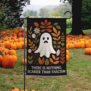 Może przedstawiać: Czarna flaga ogrodowa na Halloween z białą ilustracją ducha i napisem "THERE IS NOTHING SCARIER THAN FASCISM". Flaga jest ozdobiona jesiennymi liśćmi i kwiatami. W tle dynie.