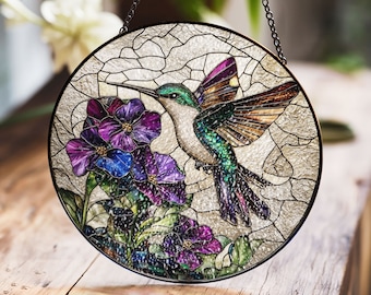 May include: A stained glass window hanging features a hummingbird with iridescent green and purple wings, hovering near purple and blue flowers. The window is round and has a black metal frame.