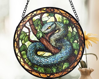May include: A stained glass window hanging depicts a blue snake with yellow eyes coiled around a tree branch. The snake is surrounded by green leaves and a brown tree trunk. The window is framed in black metal.