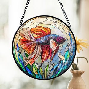 May include: A stained glass window hanging with a colorful betta fish swimming in a blue and green aquatic scene. The fish has red, orange, blue, and purple scales.