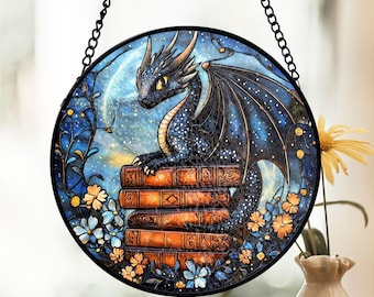 May include: A stained glass suncatcher featuring a blue dragon with a starry pattern on its wings, perched on a stack of books. The dragon is surrounded by flowers and a crescent moon.