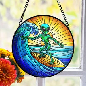 May include: Round stained glass window art featuring a green alien surfing a wave. The artwork has a black border and a chain for hanging. The background depicts a sunset with yellow and orange hues.