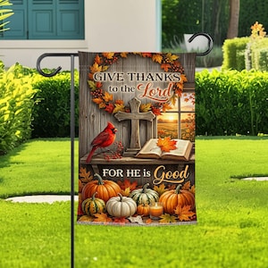 Op de afbeelding: Een rechthoekige tuinvlag met een houten achtergrond, een krans, een kruis, een open boek en pompoenen. De tekst luidt "GIVE THANKS to the Lord" en "FOR HE IS Good". Een rode kardinaal zit links en een brandende kaars staat tussen de pompoenen.