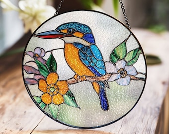 Op de afbeelding: Een hangend glas-in-loodraam met een kleurrijke ijsvogel die op een tak zit met witte en gele bloemen. De vogel heeft blauwe, gele en groene veren. De achtergrond is lichtblauw.