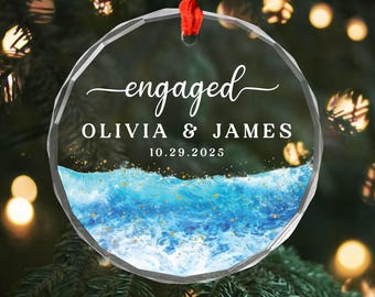 Könnte beinhalten: Ein klarer, runder Weihnachtsschmuck mit dem Wort "engaged" in eleganter Schrift. Darunter sind die Namen "OLIVIA & JAMES" und das Datum "10.29.2025" aufgedruckt. Die untere Hälfte zeigt ein blaues Wellenmuster mit goldenen Akzenten und ein rotes Band zum Aufhängen.
