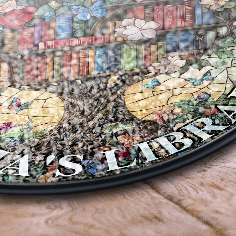 May include: A stained glass round sign with a tree design. The tree has books on the branches and butterflies flying around it. The sign says "Eliza's Library" in white lettering.