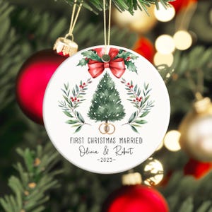 以下が含まれることがあります： お祝いのデザインが施された白いセラミック製のクリスマスオーナメント。緑のクリスマスツリー、赤いリボン、そして「FIRST CHRISTMAS MARRIED」の文字と名前、2025年が特徴です。オーナメントはクリスマスツリーに飾られています。