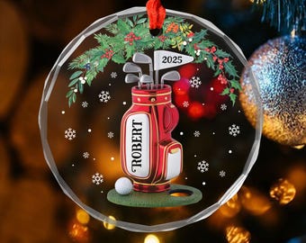 Puede incluir: Un adorno navideño redondo y transparente con una cinta roja. El adorno presenta una bolsa de golf roja con el nombre "ROBERT" y una pelota de golf sobre una superficie verde. Una bandera dice "2025". El adorno está decorado con acebo y copos de nieve.