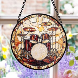 May include: A stained glass window hanging with a depiction of a drum kit. The drum kit is made of brown, yellow, and white glass. The window is round and has a black metal frame.
