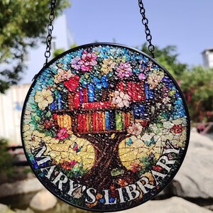 May include: A round stained glass window hanging from a black chain. The window features a colorful mosaic design of a tree with books in the branches. The text "Mary's Library" is written around the base of the tree.