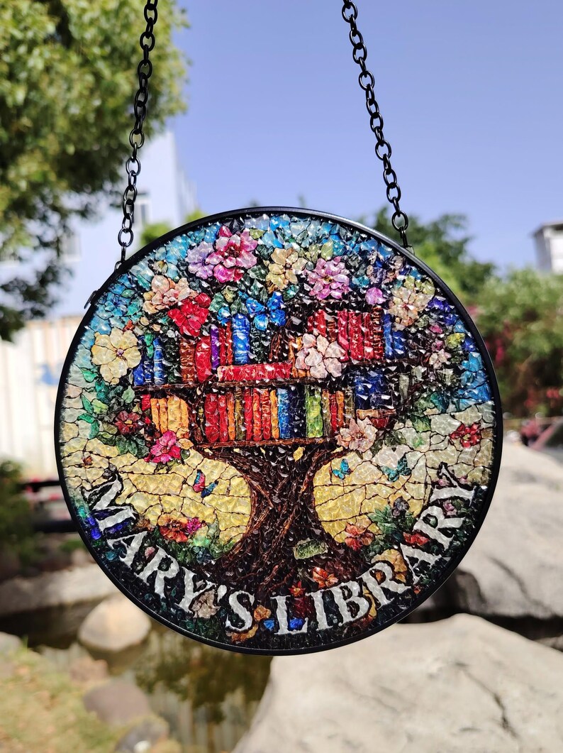 May include: A round stained glass window hanging from a black chain. The window features a colorful mosaic design of a tree with books in the branches. The text "Mary's Library" is written around the base of the tree.