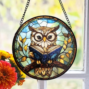 May include: A stained glass window hanging features a brown owl with large eyes holding a blue and brown book. The owl is surrounded by yellow and green leaves on a blue and white background.